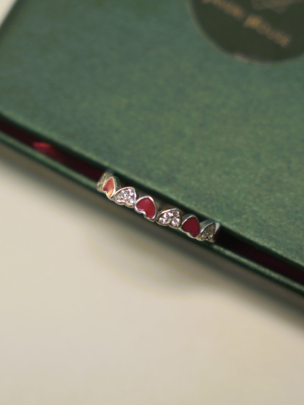 Sterling silver band ring of alternating red enamel hearts and pave set hearts, on the edge of a green box.