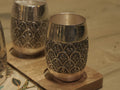 Pair of barrel shaped sterling silver drinking glasses engraved with scallop arches, on wooden coasters.