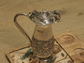 Sterling silver jug with a domed hinged lid and textured handle, standing on a wooden board.