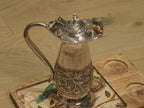 Sterling silver jug with a domed hinged lid and textured handle, standing on a wooden board.