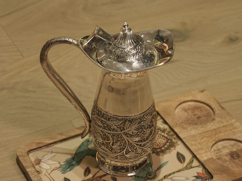 Sterling silver jug with a domed hinged lid and textured handle, standing on a wooden board.