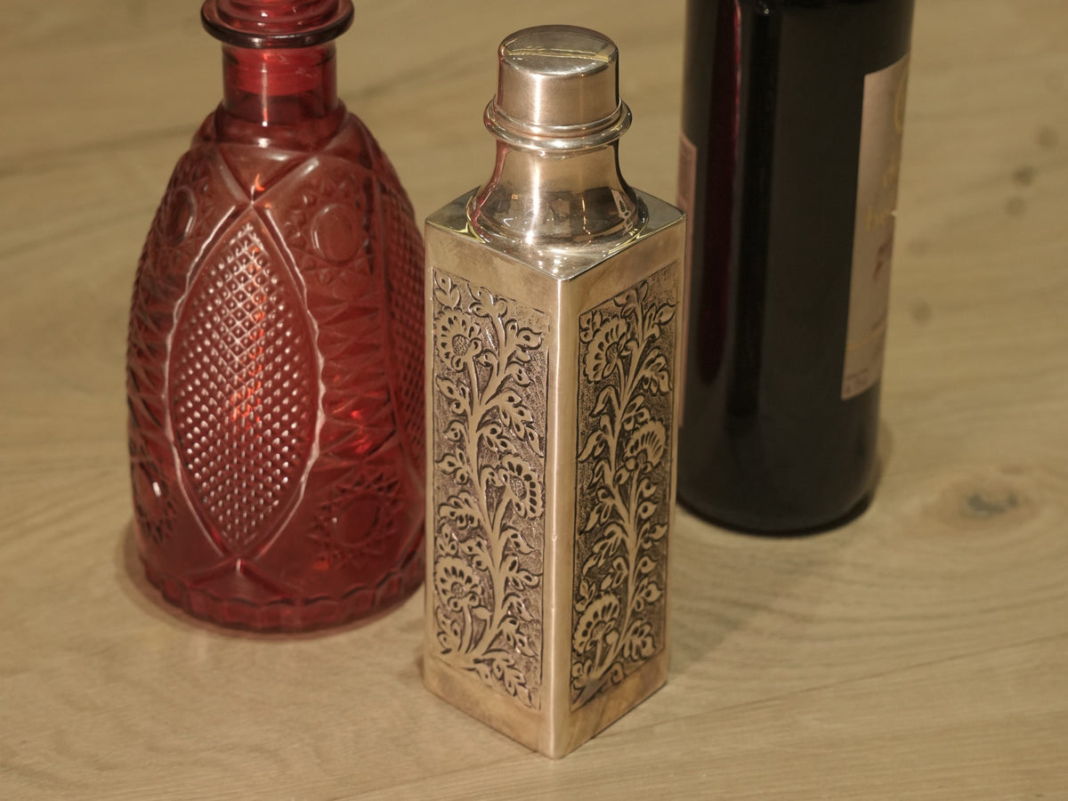 Square sterling silver water bottle with repousse floral panels and a screw cap, standing between glass bottles.