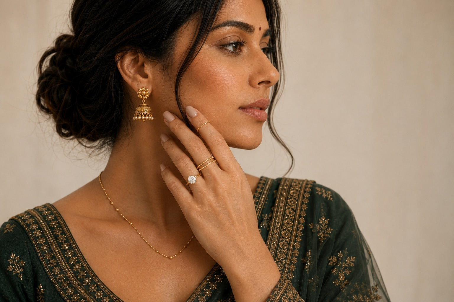A woman in profile wearing gold jewelry, including a gold jhumka earring, delicate gold rings, and a gold necklace, paired with a dark green embroidered traditional garment against a neutral beige background.